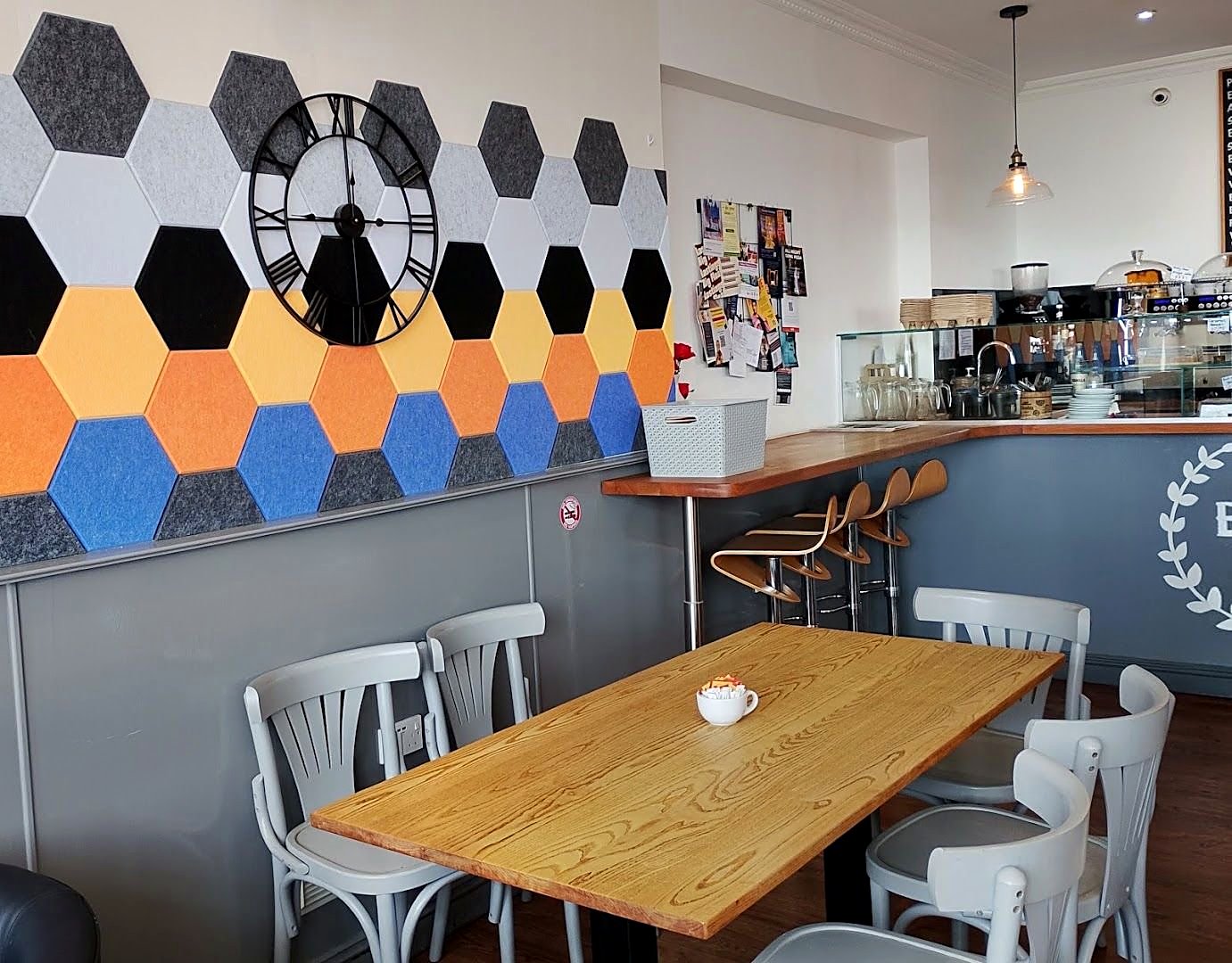 Inside Bonjour: a wall of orange, blue, grey and white hexagon panels with a large black clock, wooden tables, white bentwood chairs and the counter