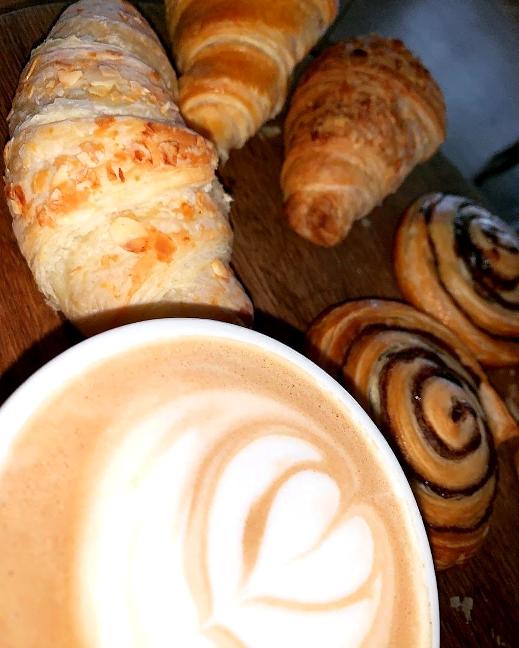 A latte with heart latte art beside a board of croissants and cinnamon swirls