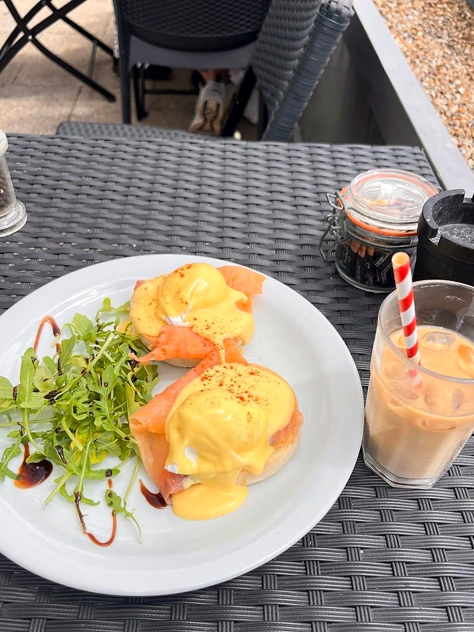 Eggs royale on a white plate at an outdoor table: smoked salmon and poached eggs with hollandaise on muffins, rocket salad, and an iced coffee with a striped straw