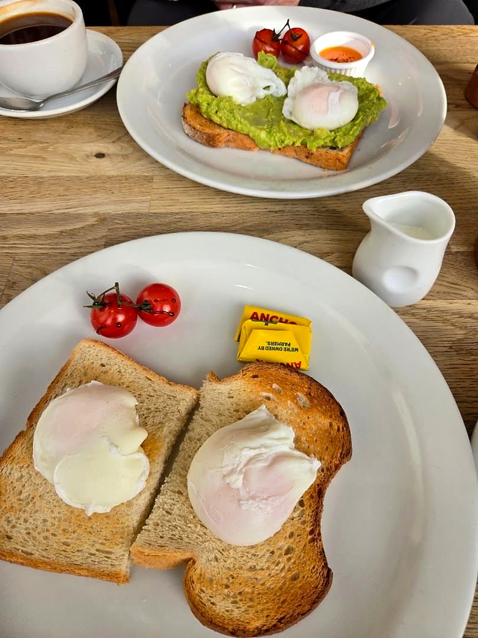 Poached eggs on toast with cherry tomatoes, and behind it eggs on smashed avocado toast, with a coffee and a jug of milk